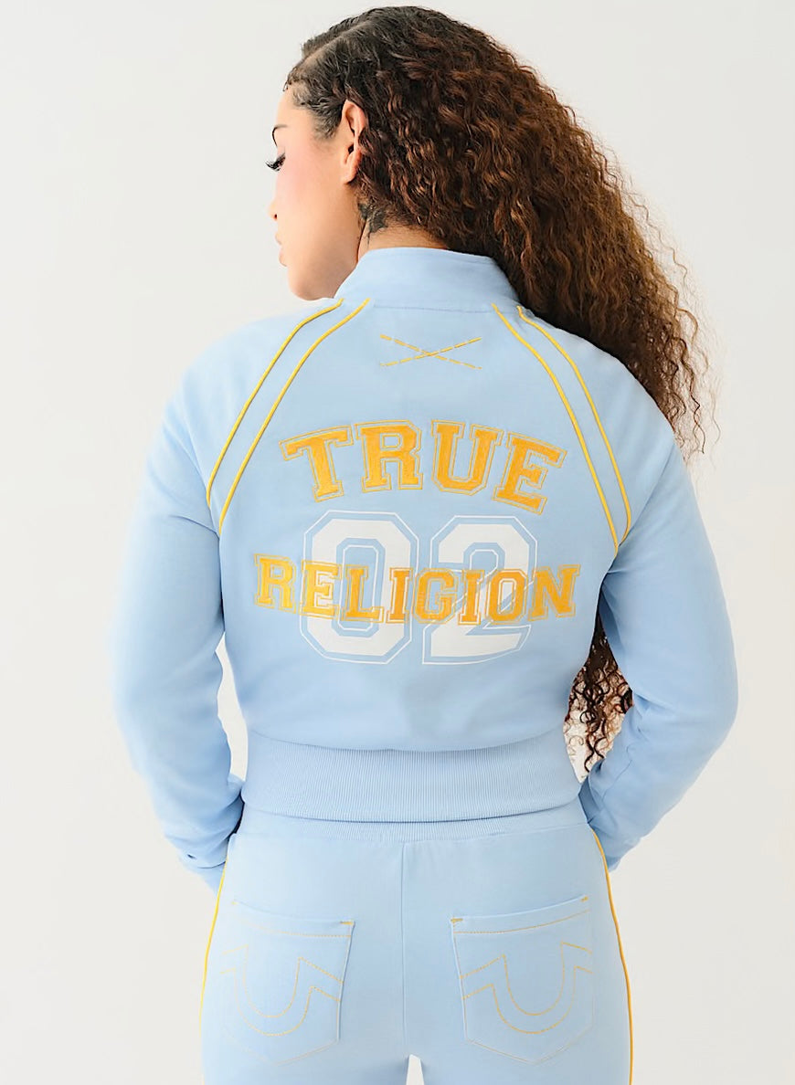 Back view of woman wearing a light blue track jacket with "TRUE RELIGION 02" in yellow and matching joggers, featuring yellow stitching.