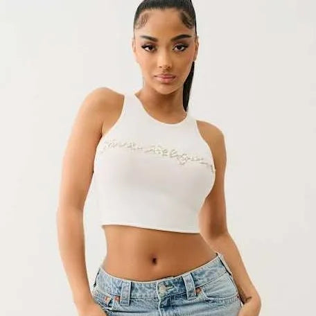 Woman wearing white cropped tank top and low-rise blue jeans, posing confidently against plain white background.