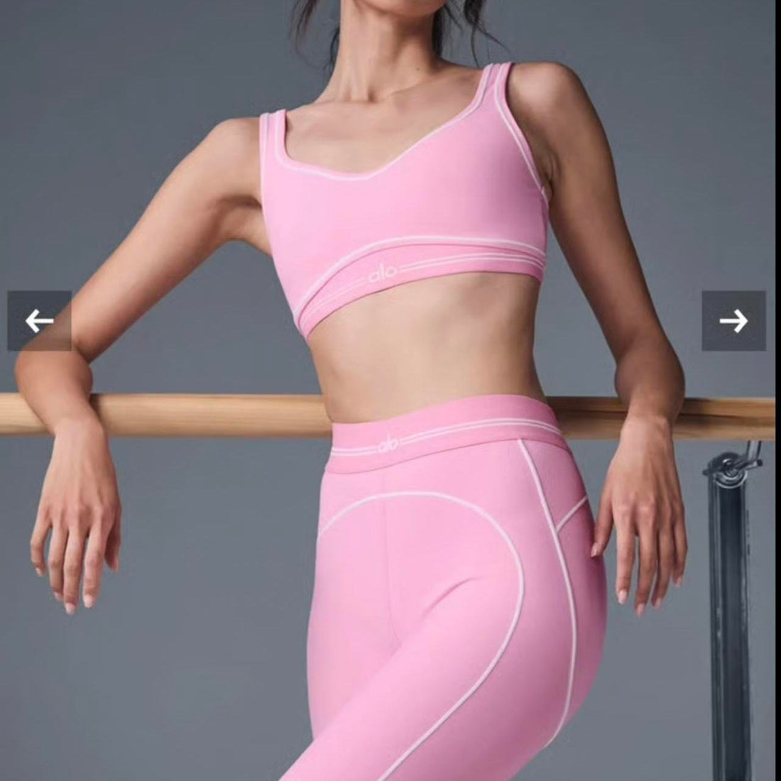 Woman wearing a matching light pink Alo Yoga sports bra and high-waisted leggings set, standing at a ballet barre with arms relaxed.