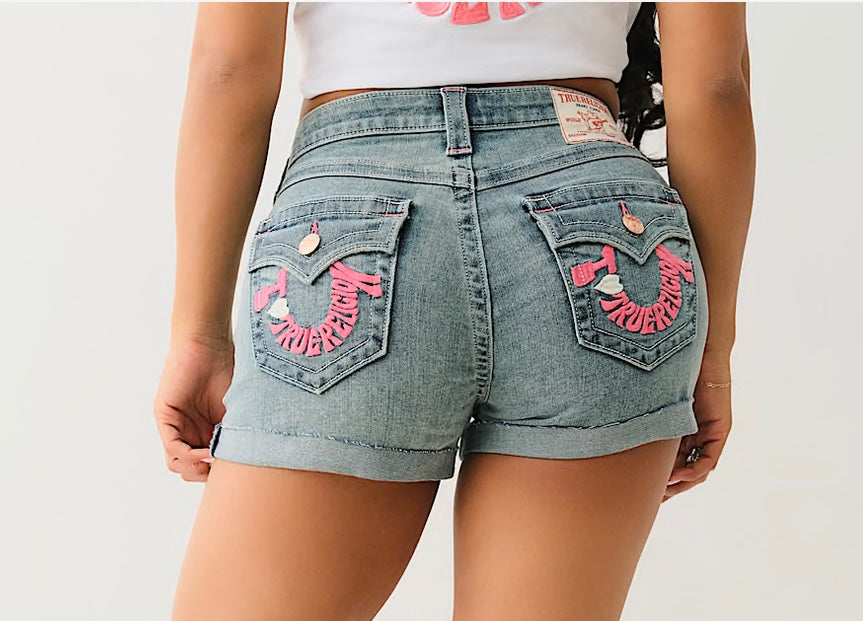 Back view of a person wearing light denim shorts with pink "True Religion" embroidery on pockets and a white cropped shirt.