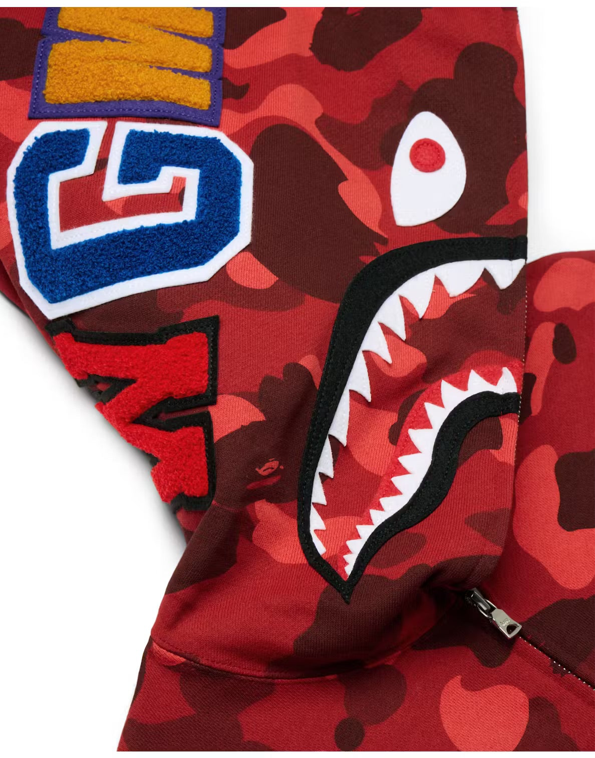 Close-up of a red BAPE hoodie hood with a shark mouth graphic, colorful patches, and a zipper at the base of the design.