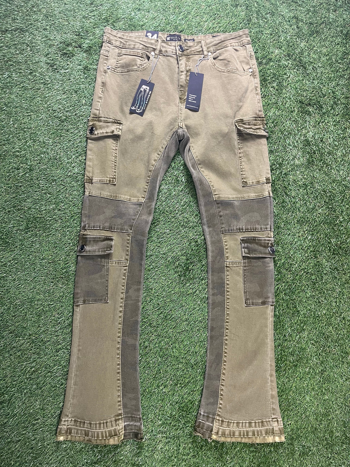 A pair of olive green cargo pants featuring side pockets, camouflage panels, and two black tags hanging from the waistband.