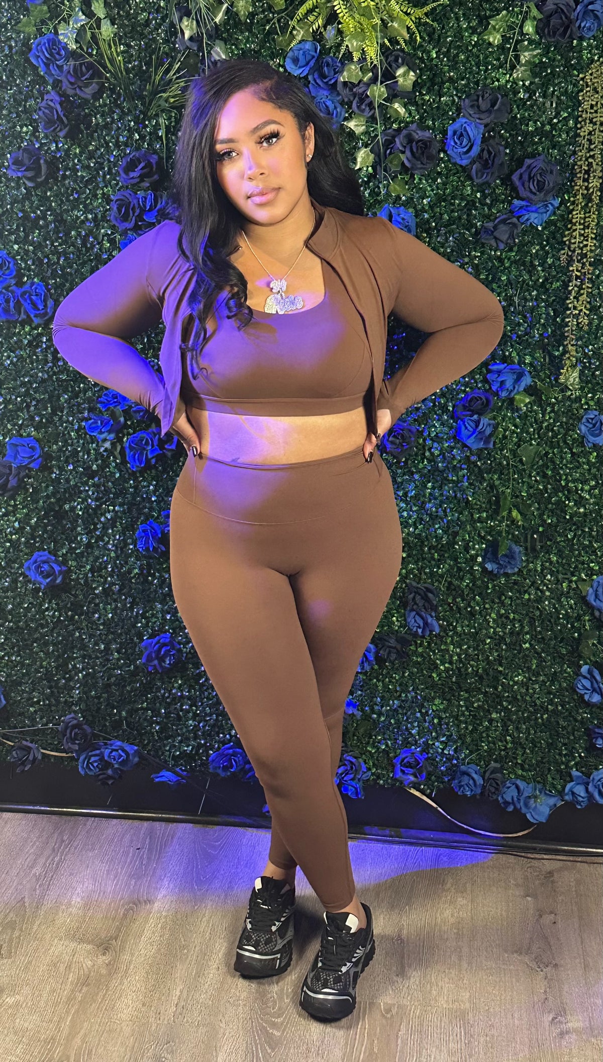 Woman in brown workout set with long sleeves and leggings, standing with hands on hips, in front of blue and green floral wall.