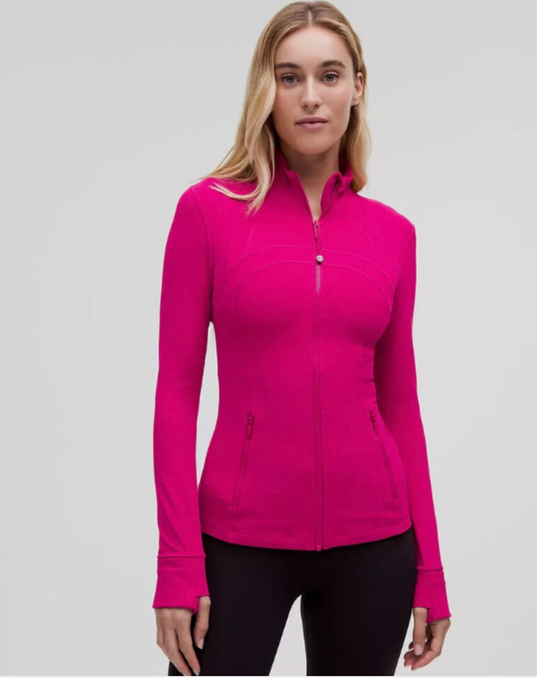 Woman wearing fitted hot pink zip-up jacket with high collar and thumbhole sleeves, paired with black leggings, standing on gray backdrop.