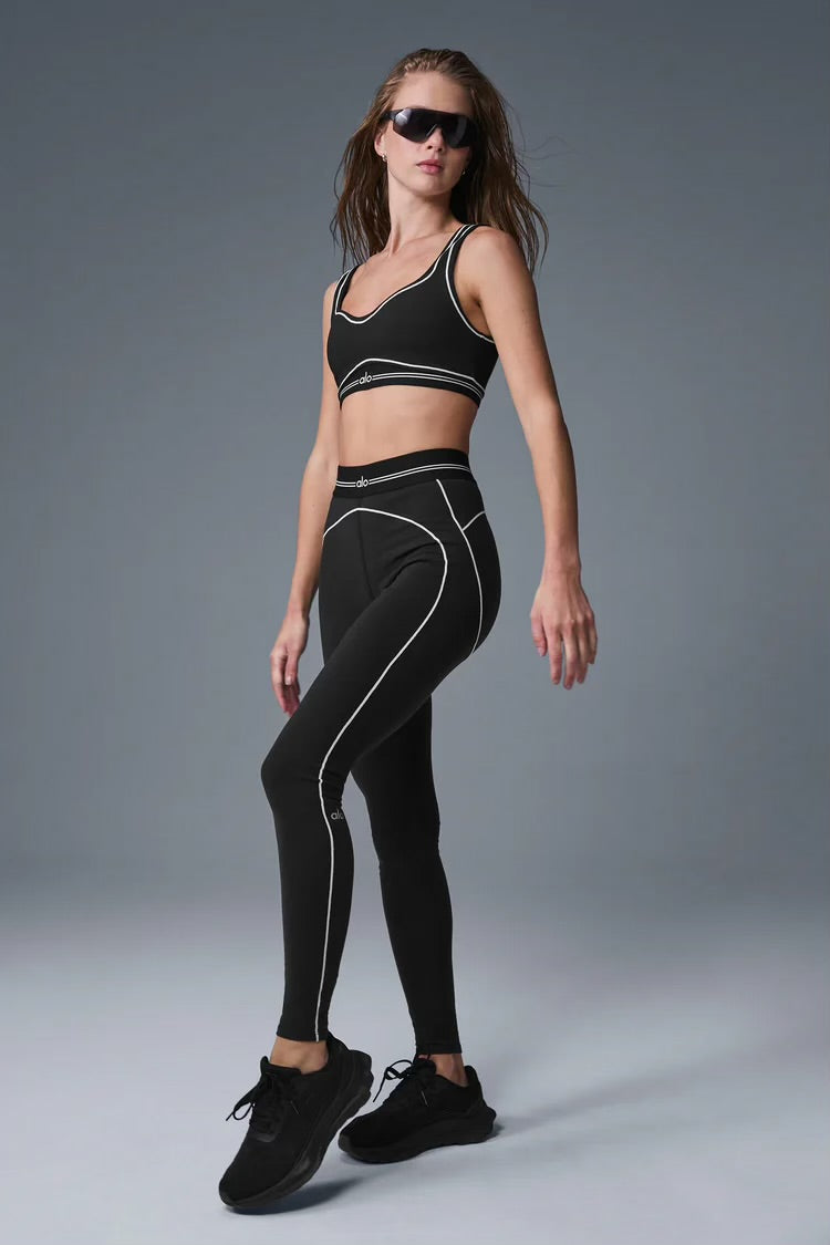 Woman wearing black sports bra and leggings with white piping, black sneakers, and sunglasses, posing on gray studio background.