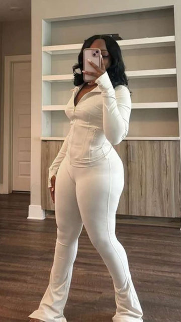A woman poses in front of a shelf, wearing a white zip-up bodysuit and matching pants, holding a phone in her left hand.