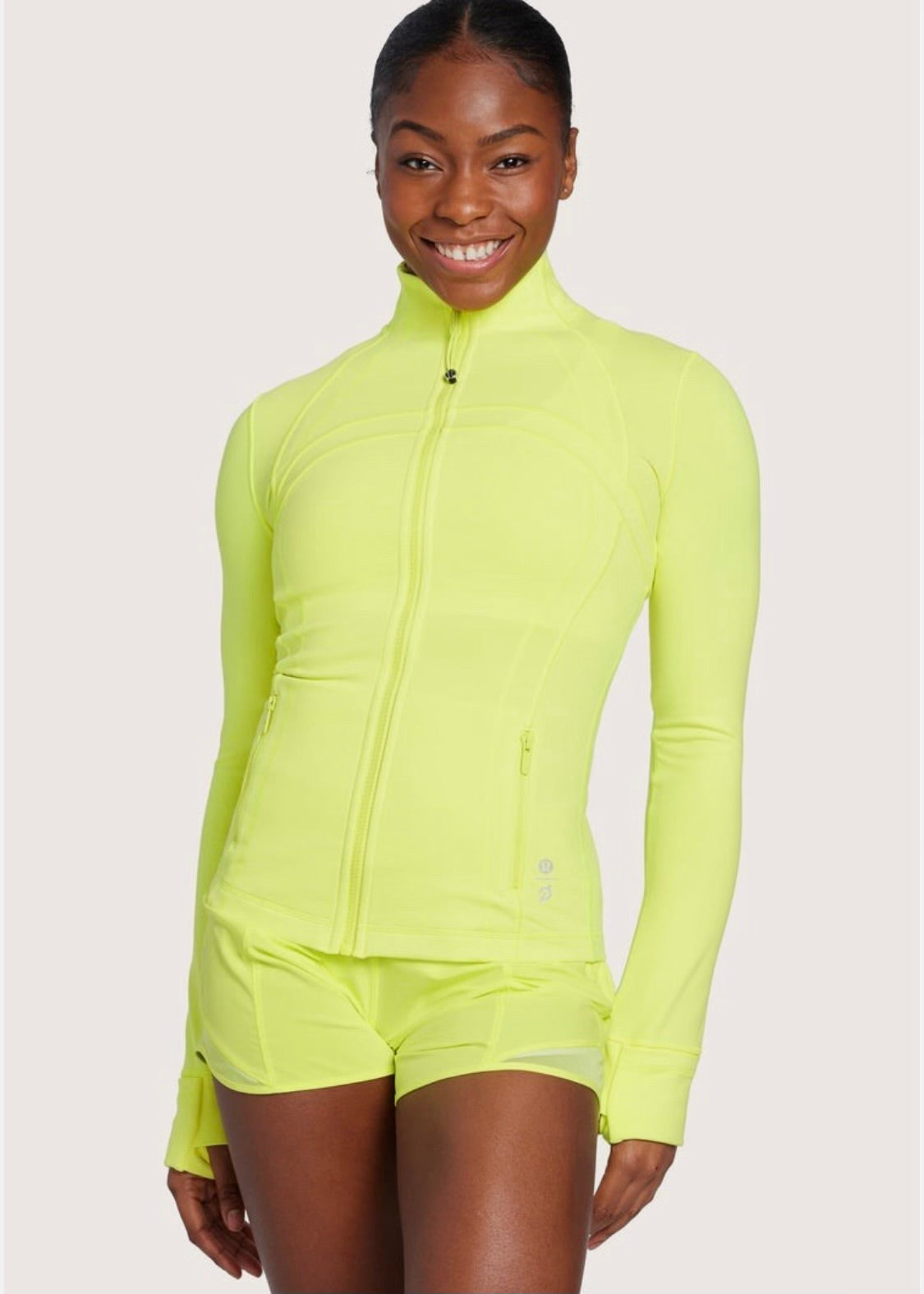 Woman smiles wearing bright neon yellow zip-up jacket and matching shorts, standing against a plain light background.
