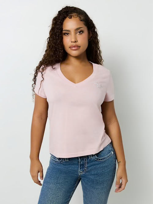 Woman with curly hair wearing a light pink V-neck T-shirt and blue jeans standing against a white background, facing forward.