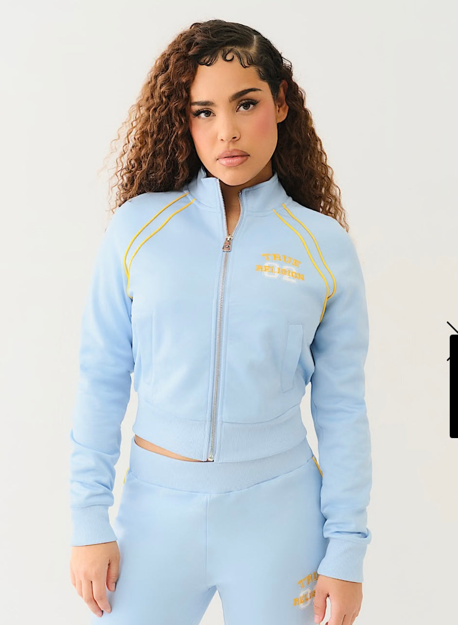 Woman wearing light blue zip-up track jacket with yellow piping and “TRUE RELIGION” text, paired with matching high-waisted joggers.