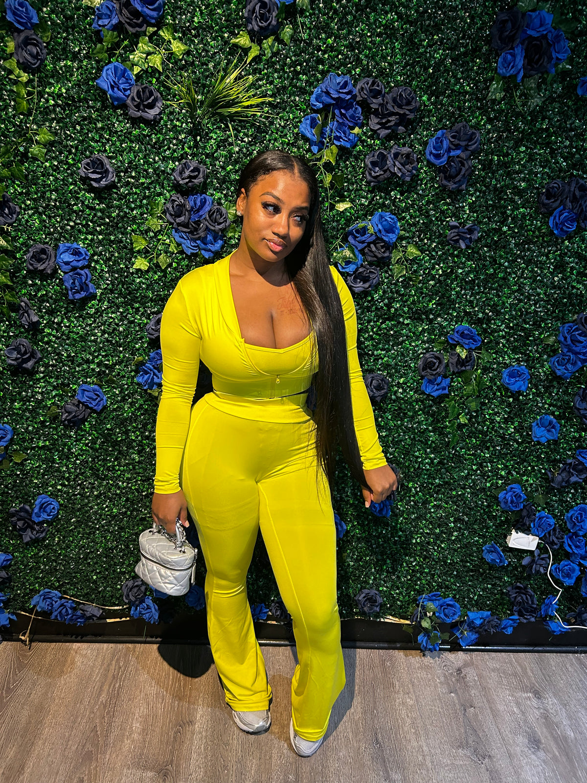 Woman in bright yellow outfit holding a white purse, standing against a green wall decorated with blue and black roses on floor.