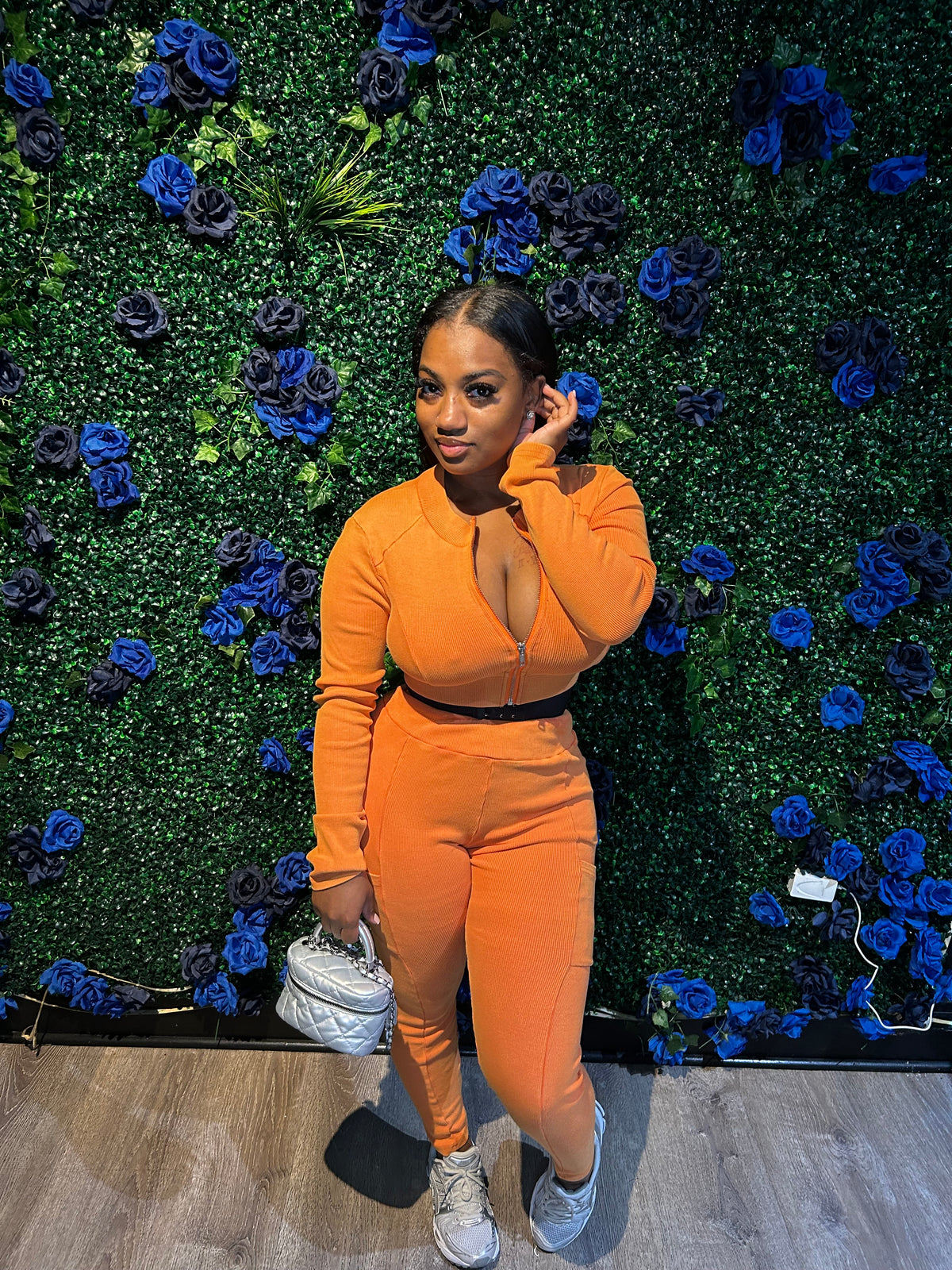 Woman in an orange zip-up two-piece outfit holding a silver quilted handbag, posing in front of a green floral wall with blue roses.