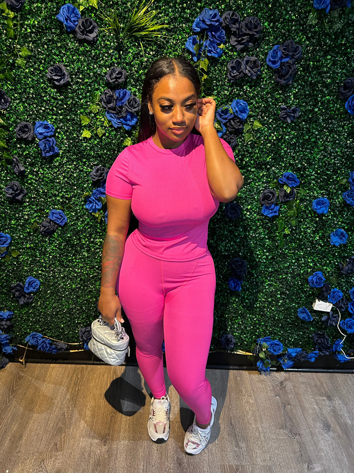 Woman wearing a pink athletic jumpsuit, holding a small silver handbag, and white sneakers, standing in front of a floral green backdrop.