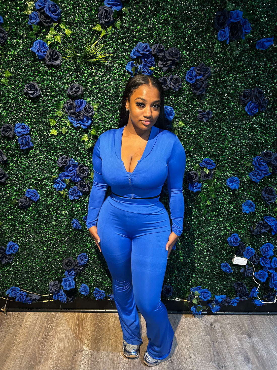 A woman wearing a blue zip-up jumpsuit with long sleeves, posing in front of a blue rose-themed wall, paired with silver sneakers.