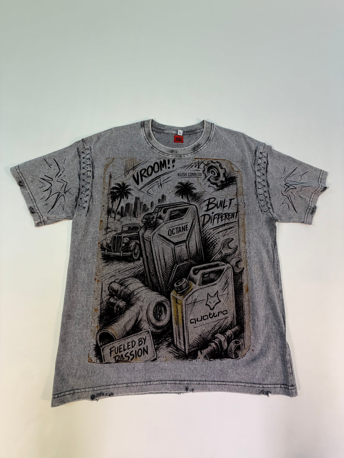 A faded gray graphic t-shirt. The shirt features a distressed, vintage-style black and white illustration of gas cans, a classic car, and palm trees, with text including 'VROOM!', 'BUILT DIFFERENT', and 'FUELED BY PASSION'.