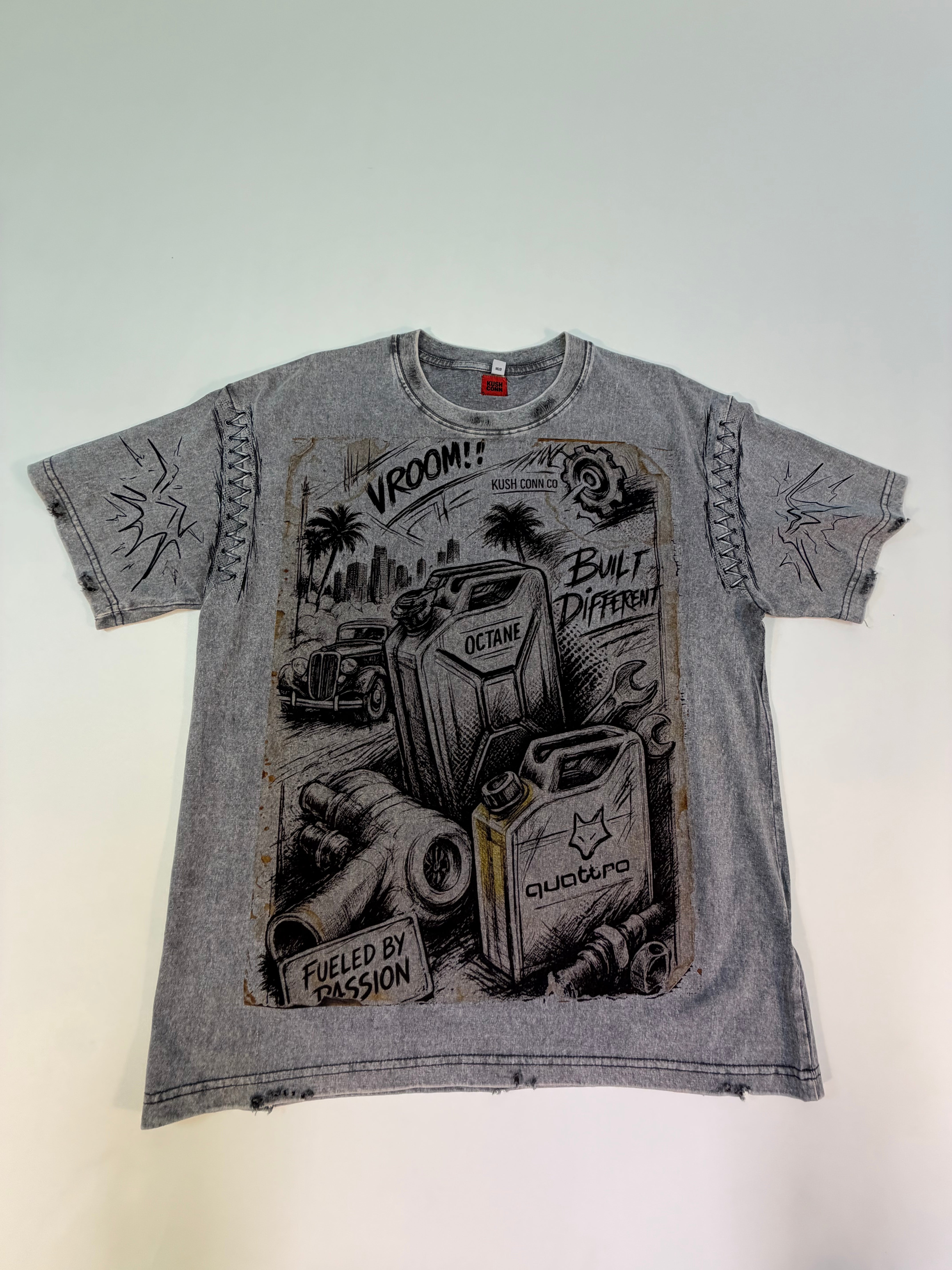 A faded gray graphic t-shirt. The shirt features a distressed, vintage-style black and white illustration of gas cans, a classic car, and palm trees, with text including 'VROOM!', 'BUILT DIFFERENT', and 'FUELED BY PASSION'.