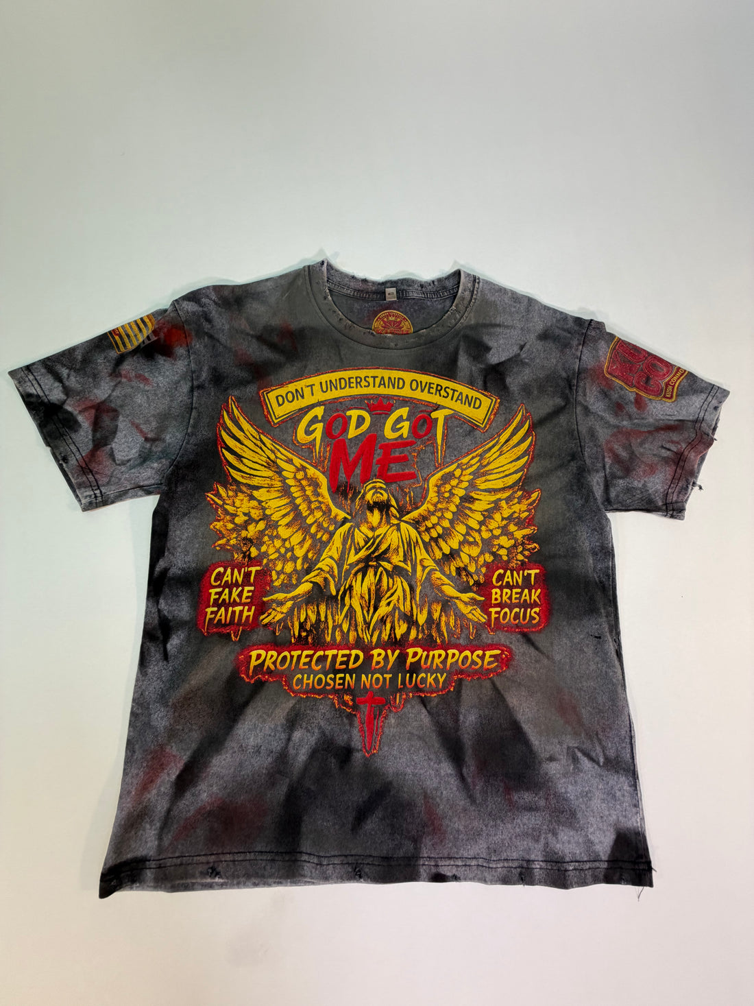 A short-sleeve graphic t-shirt with a distressed black and gray acid-wash finish, featuring a large red and yellow graphic of a winged figure and text including 'GOD GOT ME' and 'PROTECTED BY PURPOSE'.