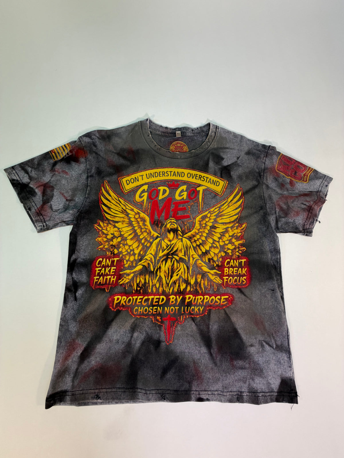A short-sleeve graphic t-shirt with a distressed black and gray acid-wash finish, featuring a large red and yellow graphic of a winged figure and text including 'GOD GOT ME' and 'PROTECTED BY PURPOSE'.